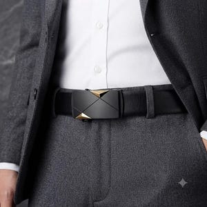 May include: A black leather belt featuring a rectangular buckle with a matte black finish and two small, triangular gold-tone metallic accents at the top and bottom corners, worn over grey trousers and a white button-down shirt.