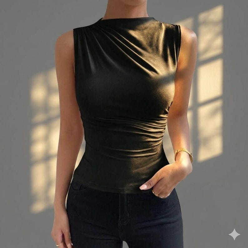 May include: A sleeveless black top featuring a high neckline and subtle draped fabric detailing across the chest. The garment is fitted through the torso and is paired with dark denim jeans, creating a sleek and streamlined silhouette.