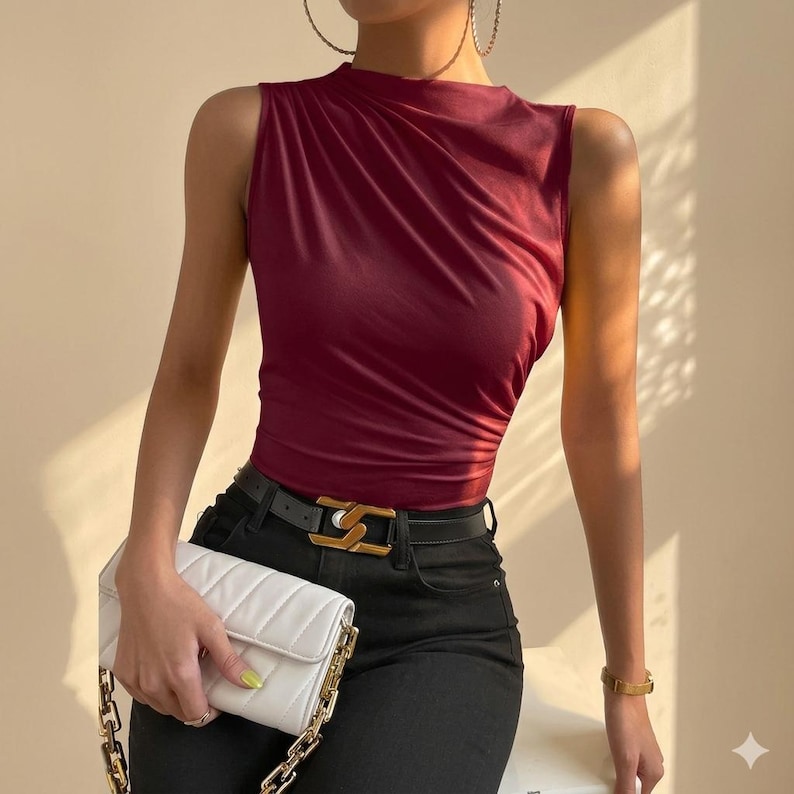 May include: A sleeveless burgundy top with a high neckline and gathered fabric detail, paired with black trousers, a black belt featuring a gold-toned buckle, and a white quilted rectangular handbag with a gold-toned chain shoulder strap.