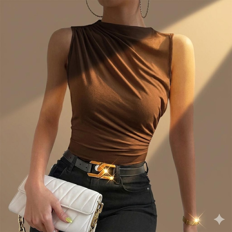 May include: A sleeveless brown top with gathered fabric detail at the shoulder, paired with black trousers featuring a black belt with a gold-toned buckle, and a white quilted handbag with a gold-toned chain strap and gold-toned hardware.