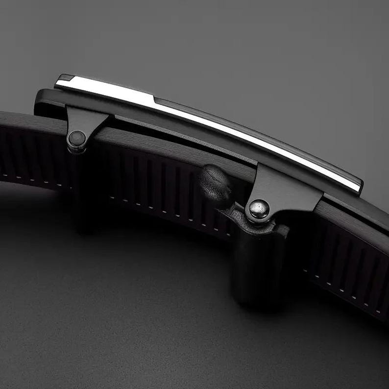 May include: A black belt buckle featuring a matte finish with a sleek silver-toned metallic accent strip along the top edge. The buckle mechanism includes a hinged lever and a ridged interior structure designed to grip the belt strap securely.
