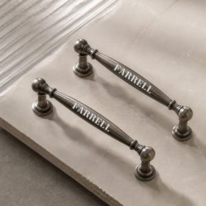 Könnte beinhalten: Zwei Schubladengriffe aus Metall im Antik-Stil mit dunkler, verwitterter Oberfläche. Jeder Griff hat ein abgerundetes, konisches Design mit kugelförmigen Enden und dem Wort FARRELL, das in weißen Großbuchstaben in der Mitte aufgedruckt ist.