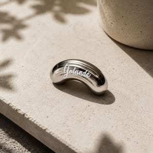 Può includere: Un oggetto metallico a forma di mezzaluna, di colore argento lucido con una superficie liscia e riflettente. Il nome Yolande è inciso in corsivo bianco al centro del corpo curvo, caratterizzato da sottili scanalature orizzontali.