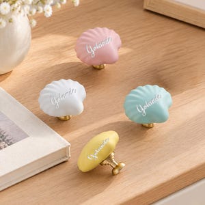 Puede incluir: Cuatro pomos para armario con forma de concha en colores rosa pastel, blanco, azul claro y amarillo. Cada pomo tiene una base de metal dorado y el nombre Yolanda grabado en letra cursiva en la parte frontal del tirador.