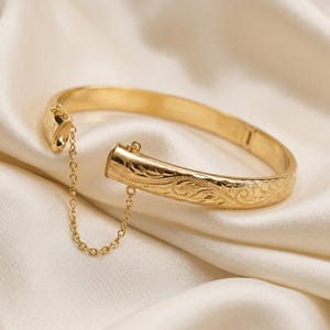 May include: A gold-toned hinged bangle bracelet featuring an intricate engraved floral pattern along the outer surface. The bracelet includes a delicate gold-toned safety chain connecting the two ends, which are designed to open and close securely.