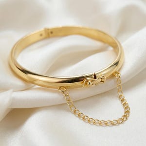 May include: A polished gold-tone hinged bangle bracelet featuring a smooth, rounded band and a secure box clasp closure. A delicate, thin gold-tone chain is attached to both sides of the clasp, creating a draped link accent across the front.