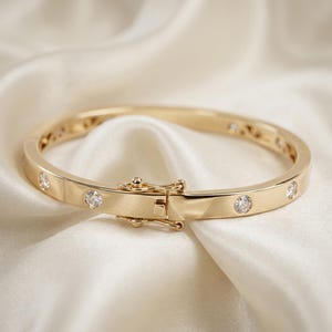 May include: A gold-toned hinged bangle bracelet featuring a polished finish and multiple round, clear, faceted stones set at intervals along the band. The bracelet includes a secure, integrated box clasp mechanism with a side safety latch.