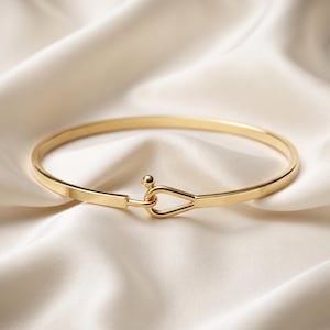 May include: A gold-toned metal bangle bracelet featuring a smooth, polished finish. The bracelet has a circular band that connects at the front with a hook-and-eye style closure, consisting of a small spherical knob and a teardrop-shaped loop.