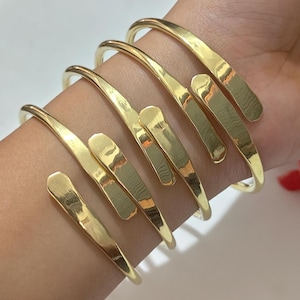 May include: A set of three gold-toned metal cuff bracelets, each featuring a smooth, polished finish and a bypass design with two flat, rounded ends that overlap slightly. The bracelets are worn stacked together on a wrist, showcasing a reflective surface.