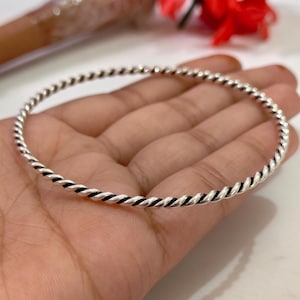 Può includere: Un bracciale rigido circolare e sottile realizzato in metallo lucido color argento. La superficie del bracciale presenta un motivo a corda ritorta continuo e ripetitivo che crea un aspetto a spirale testurizzato lungo la circonferenza.