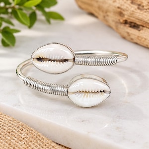 Puede incluir: Una pulsera abierta de tono plateado con dos conchas de cauri blancas en cada extremo. La banda metálica presenta un diseño de alambre enrollado que conduce a las conchas, creando un aspecto simétrico para este accesorio de muñeca.
