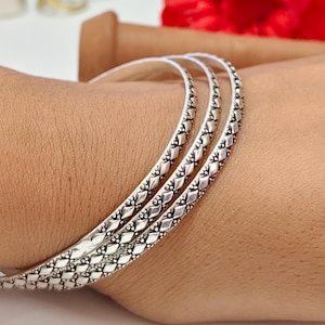 May include: A set of three silver-toned bangles featuring a repeating diamond-shaped pattern with small dot accents. The metal surface has a darkened finish within the recessed areas to highlight the intricate geometric texture of the jewelry.
