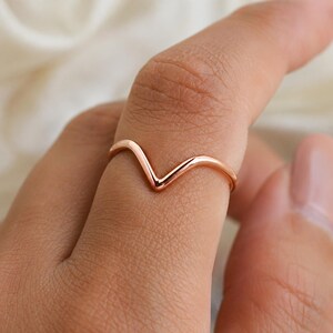 Könnte beinhalten: Ein polierter Ring aus Roségold mit einem schlichten, V-förmigen Chevron-Design. Die Ringschiene hat ein glattes, abgerundetes Profil und eine durchgehende metallische Oberfläche, was für ein klares und schlichtes Erscheinungsbild sorgt.