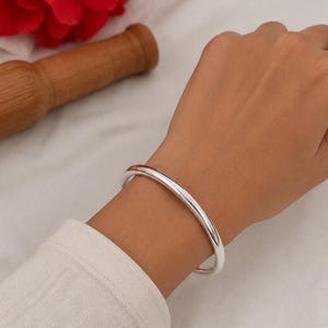 May include: A smooth, polished silver bangle bracelet with a rounded profile. The bracelet features a continuous, solid band design with a high-shine reflective surface that catches the light, encircling the wrist with a clean and simple aesthetic.