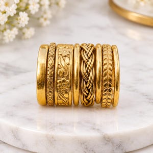 May include: A set of eight gold-toned metal rings arranged in a row. The collection includes bands with smooth surfaces, intricate floral engravings, braided textures, and geometric patterns, all featuring a consistent metallic yellow finish.