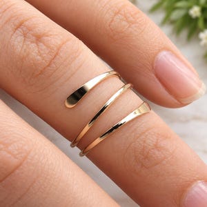 May include: A gold-toned bypass ring featuring a triple-wrap design with smooth, polished surfaces. The ring consists of three continuous, slender bands that spiral around the finger, creating a layered appearance with rounded, tapered ends.