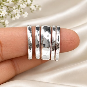 May include: A set of five silver-toned rings featuring a hammered, textured finish. The collection includes four slender bands and one wider central band, all displaying a reflective, metallic surface with irregular indentations.