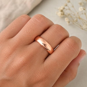 May include: A polished rose gold band ring with a smooth, rounded surface and a high-shine metallic finish. The ring features a simple, solid construction with a consistent width throughout the entire circumference of the circular band.