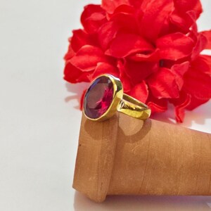 Peut inclure: Une bague en or ornée d'une grande pierre précieuse ovale rouge foncé montée sur un anneau en métal lisse. La bague est posée sur un objet cylindrique brun clair texturé, avec une fleur rouge vive et frisée visible en arrière-plan.