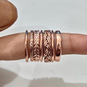 Pode incluir: Um conjunto de sete anéis em tom de cobre com várias texturas e designs, incluindo bandas lisas, um padrão trançado, um motivo de folha e uma gravação geométrica em forma de diamante, dispostos lado a lado num dedo.
