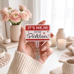 Può includere: Un porta badge rettangolare con sfondo bianco, bordo rosso e il testo IT'S ME, HI I'M THE Problem in caratteri neri e rossi, decorato con piccole icone di stelle e cuori rossi, fissato a un cinturino in plastica trasparente e clip metallica.