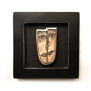 May include: A framed artwork featuring a ceramic sculpture of two faces, one on each side of the piece. The faces are stylized and have simple features, with black lines for eyes, nose, and mouth. The sculpture is a light brown color and is set against a black background.