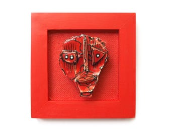 May include: A red framed artwork featuring a stylized face sculpture. The face is red with black outlines and white accents, mounted on a red fabric background. The frame is square and painted red.