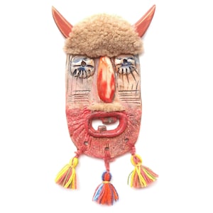 Puede incluir: Una máscara artesanal y caprichosa con una parte superior de piel marrón claro y cuernos rojos. La cara está pintada con detalles rojos, azules y blancos, y tiene una boca grande y abierta. Borlas coloridas cuelgan de la parte inferior. Una pieza decorativa única.