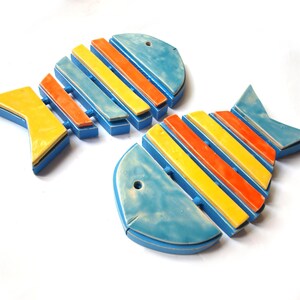 Könnte beinhalten: Zwei Keramik-Fisch-Wanddekorationen, eine blau und gelb, die andere blau und orange, mit gelben und orangen Streifen.