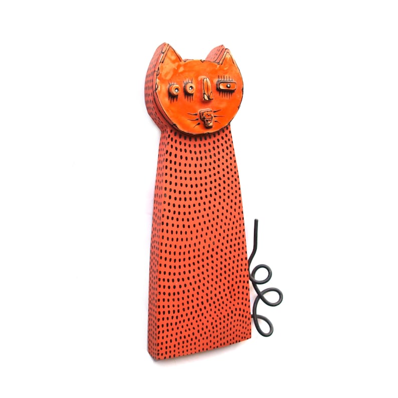 May include: Orange cat sculpture with black dot pattern and a whimsical face. The cat has a long, rectangular body and a curled tail. The sculpture is made of a textured material.