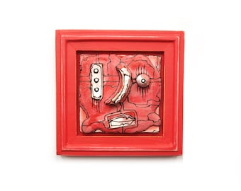 May include: A red framed artwork with a colorful abstract face design. The face is made of textured materials and has a red, white, and black color scheme.