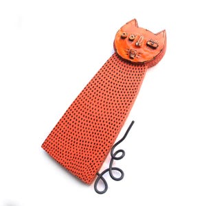 May include: Orange cat-shaped decorative item with black dot pattern on the body and a curled tail. The cat's face has detailed features, including eyes, nose, and mouth. The item is set against a white background.