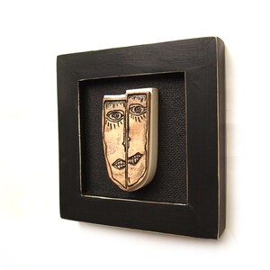 May include: A framed artwork featuring two ceramic faces, one with a light brown glaze and the other with a silver glaze. The faces are stylized and have simple black lines for features. The artwork is framed in a black wood frame.