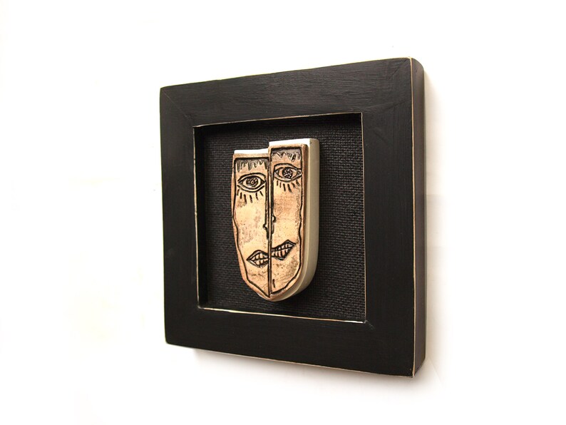 May include: A framed artwork featuring two ceramic faces, one with a light brown glaze and the other with a silver glaze. The faces are stylized and have simple black lines for features. The artwork is framed in a black wood frame.