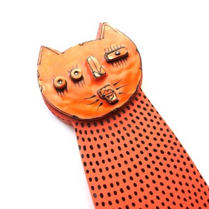 May include: Orange ceramic cat sculpture with black dot pattern. The cat has a whimsical design with detailed facial features, including eyes, nose, and mouth. The sculpture is set against a white background.