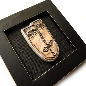 May include: A framed ceramic wall art piece featuring two stylized faces with black lines and brown paint. The faces are split in half and joined together in the middle.