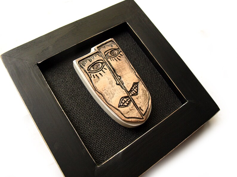 May include: A framed ceramic wall art piece featuring two stylized faces with black lines and brown paint. The faces are split in half and joined together in the middle.