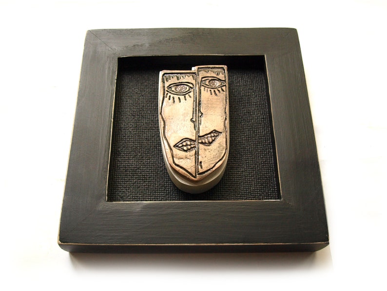 May include: A framed artwork featuring a ceramic sculpture of two faces, one on each side of the piece. The faces are stylized and have a copper-colored glaze. The frame is black with a gold-colored inner edge.
