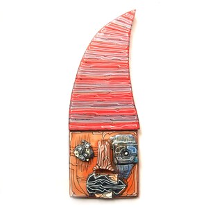 May include: Abstract art piece with a red, orange, and blue color scheme. The artwork features a tall, curved red top with horizontal lines. The lower section has a face-like design with geometric shapes and patterns. The piece is mounted on a white background.