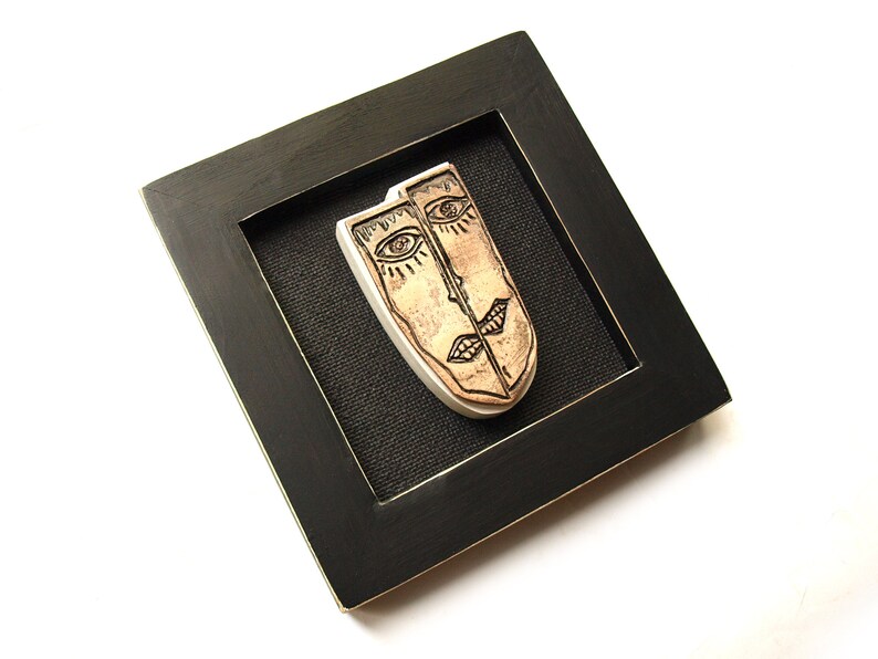 May include: A framed ceramic wall art piece featuring two stylized faces with black lines and brown glaze. The faces are joined together and have a black background.