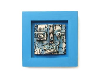 May include: A square wall art piece featuring a light blue wooden frame surrounding a ceramic tile. The tile displays a stylized face with black outlines, light blue and white tones, a raised square eye, a round eye, and a layered mouth section.