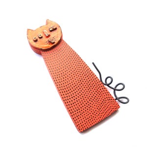 May include: Orange cat-shaped wall art with black dot pattern and a curled black metal tail. The cat's face has detailed features, including eyes, nose, and mouth. The art piece is set against a white background.
