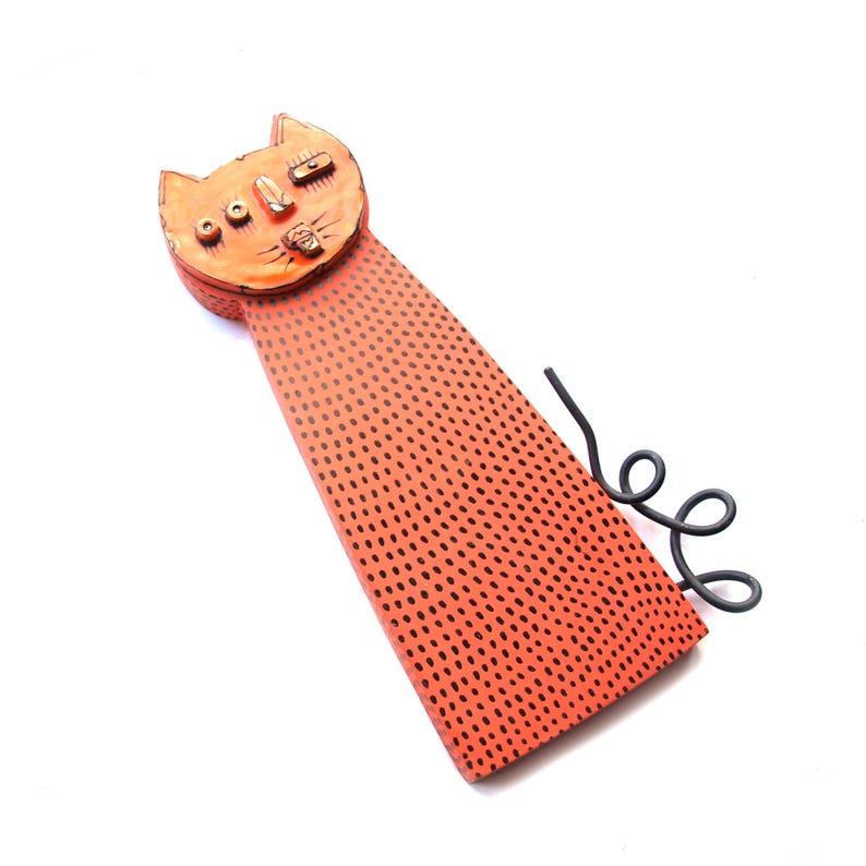 May include: Orange cat-shaped wall art with black dot pattern and a curled black metal tail. The cat's face has detailed features, including eyes, nose, and mouth. The art piece is set against a white background.