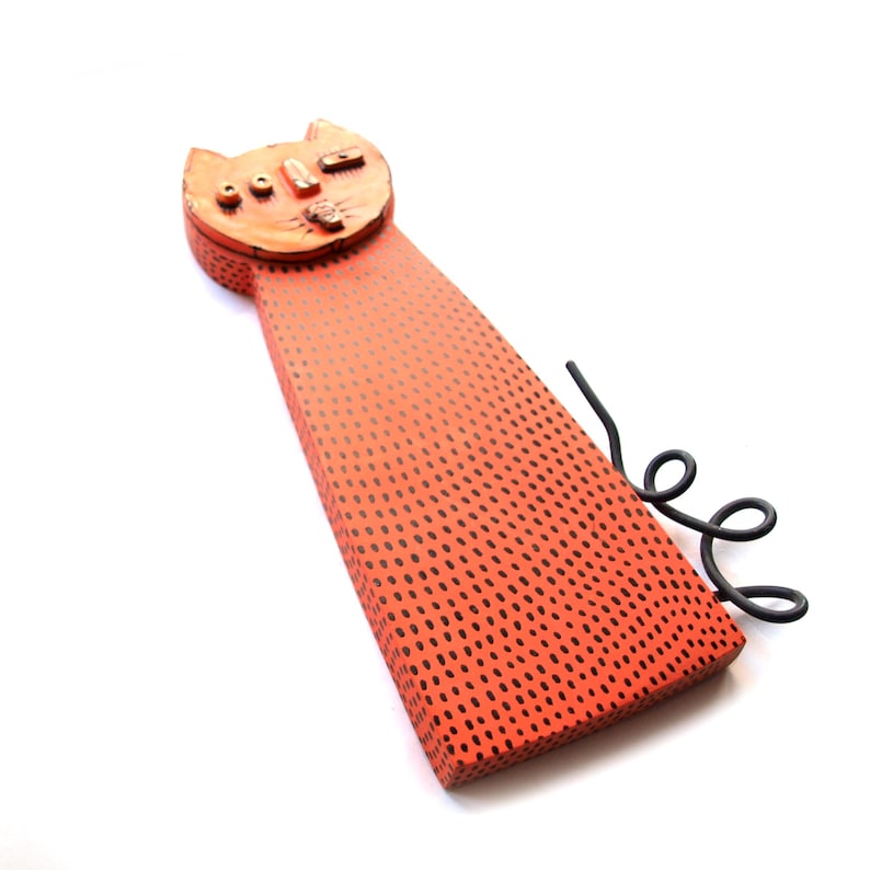 May include: Orange cat-shaped decorative object with black dotted pattern and a curled black wire tail. The cat's head has simple facial features. The object is set against a white background.