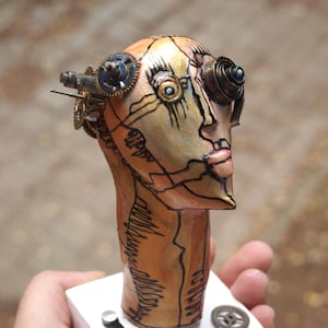 Może przedstawiać: Rzeźba głowy w stylu steampunk, w odcieniach pomarańczu i beżu. Posiada szczegółową czarną grafikę liniową, zdobienia w kształcie kół zębatych i mechaniczne oko. Rzeźba jest trzymana w dłoni, ukazując jej skomplikowany design.