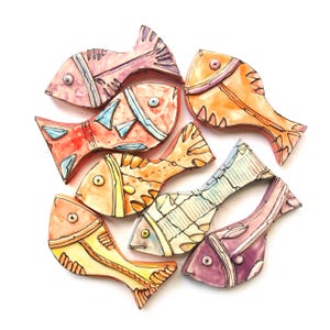 Puede incluir: Colección de peces de cerámica pintados a mano en varios colores, incluyendo rosa, naranja, amarillo y azul. Cada pez tiene patrones y detalles únicos, con un acabado brillante. Los peces están dispuestos en un patrón circular sobre un fondo blanco.