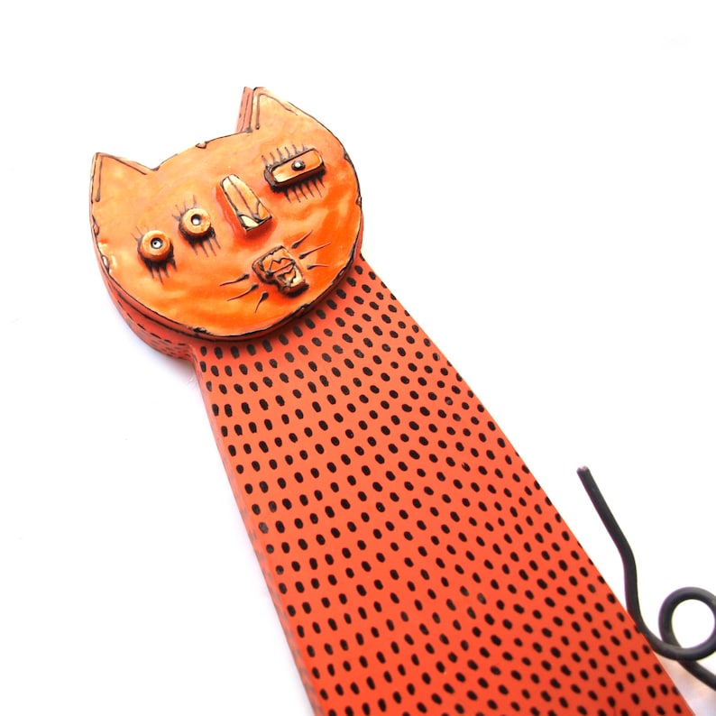 May include: Orange cat-shaped wall decor with a long, dotted body and a whimsical face. The cat's head features button-like eyes and a textured nose and mouth. The body is a solid orange with evenly spaced black dots. A black metal tail curls at the end.