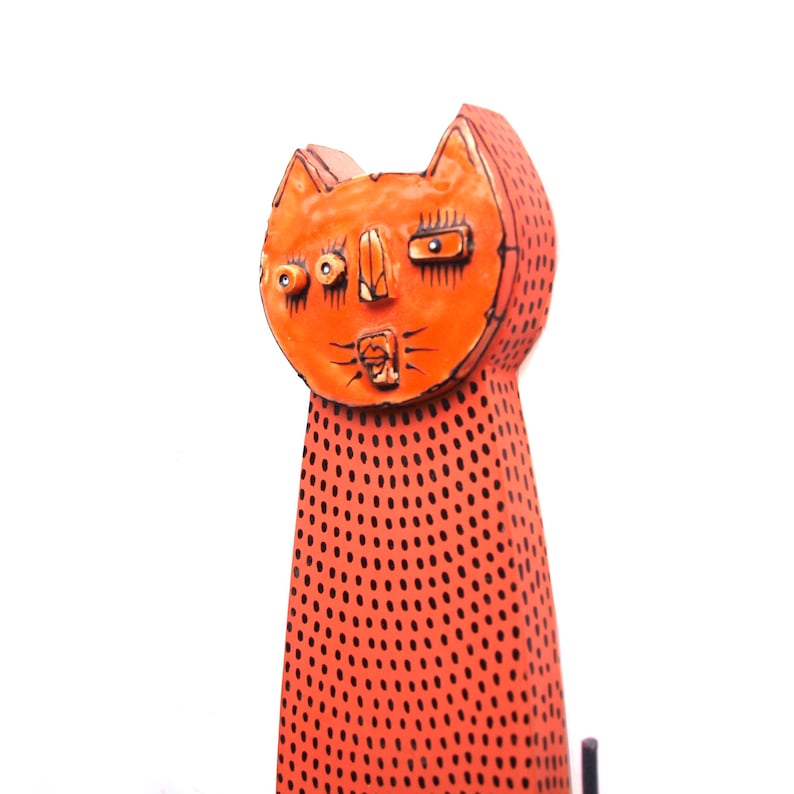May include: Orange ceramic cat figurine with black dot pattern. The cat has a stylized face with large eyes and a surprised expression. The ears are pointed, and the body is elongated.