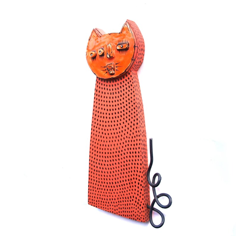 May include: A whimsical orange cat sculpture with black dot detailing. The cat has a stylized face and a curled tail. The sculpture is made of a solid material and is suitable for indoor or outdoor display.