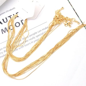 Puede incluir: Un collar de cadenas múltiples en tono dorado con eslabones finos y delicados y un cierre de mosquetón. El collar está sujeto a una tarjeta de presentación blanca con el texto impreso "by Queen Elizabeth" y "BEAUTIFUL MOOD".